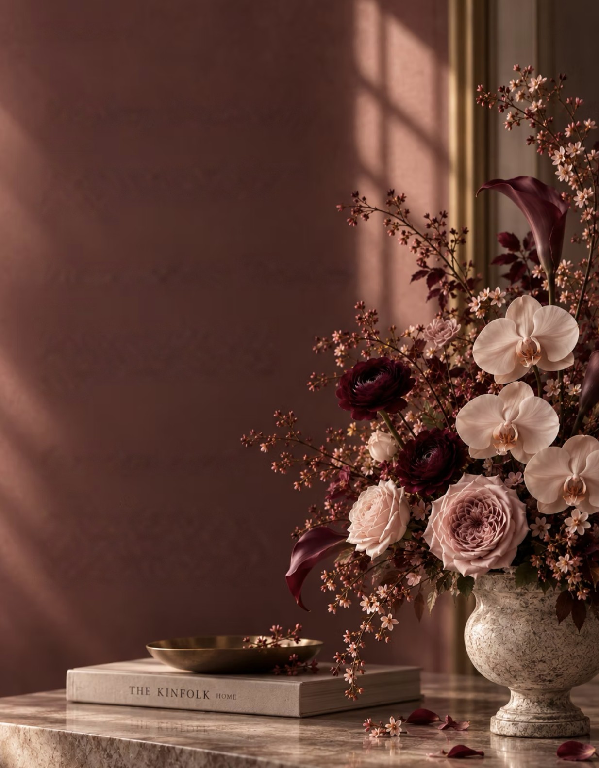 A stone urn of orchids, roses and burgundy ranunculus beside a brass dish and a closed book.