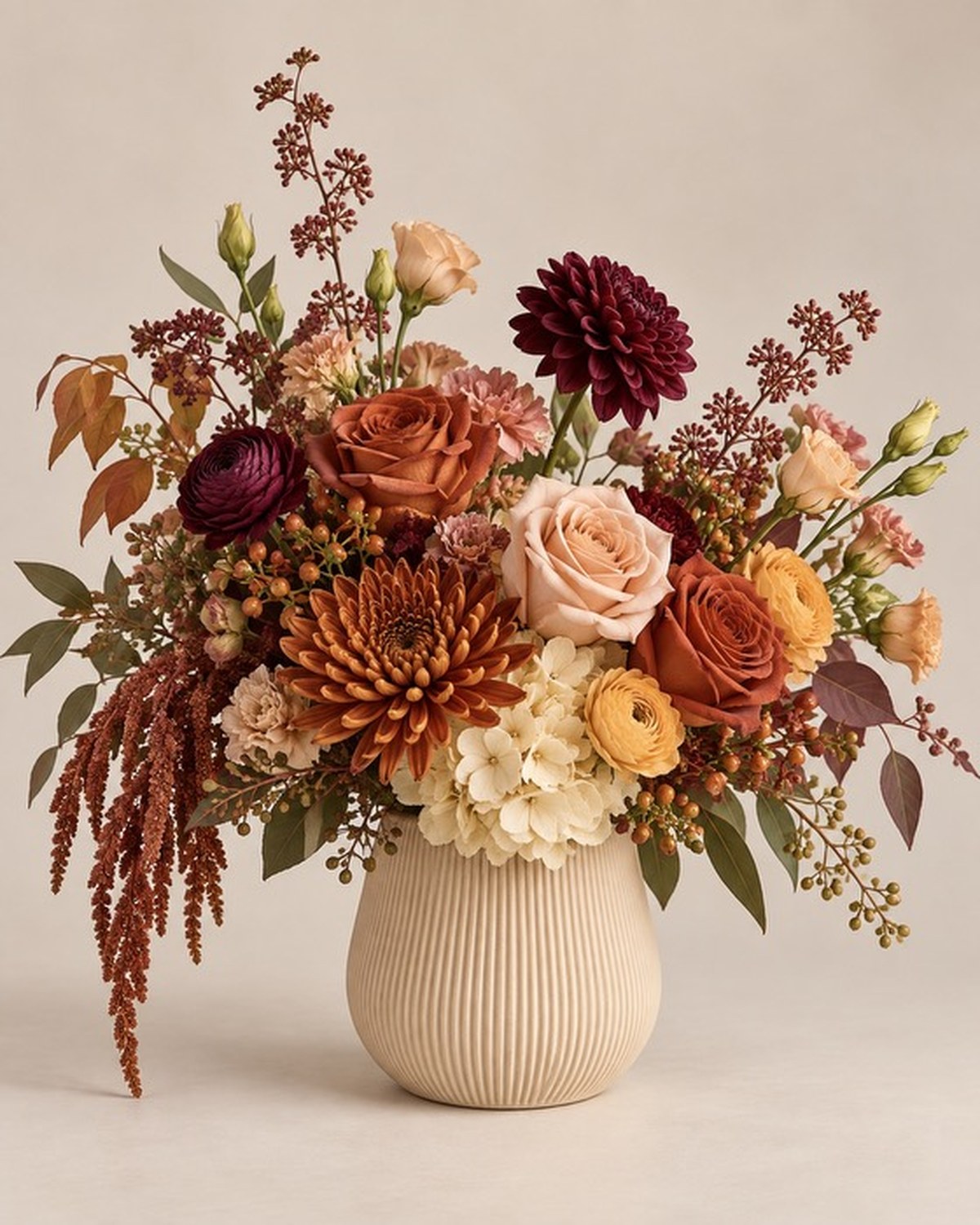 A round cream vase holding burgundy dahlias, orange roses, peach ranunculus and autumn foliage.