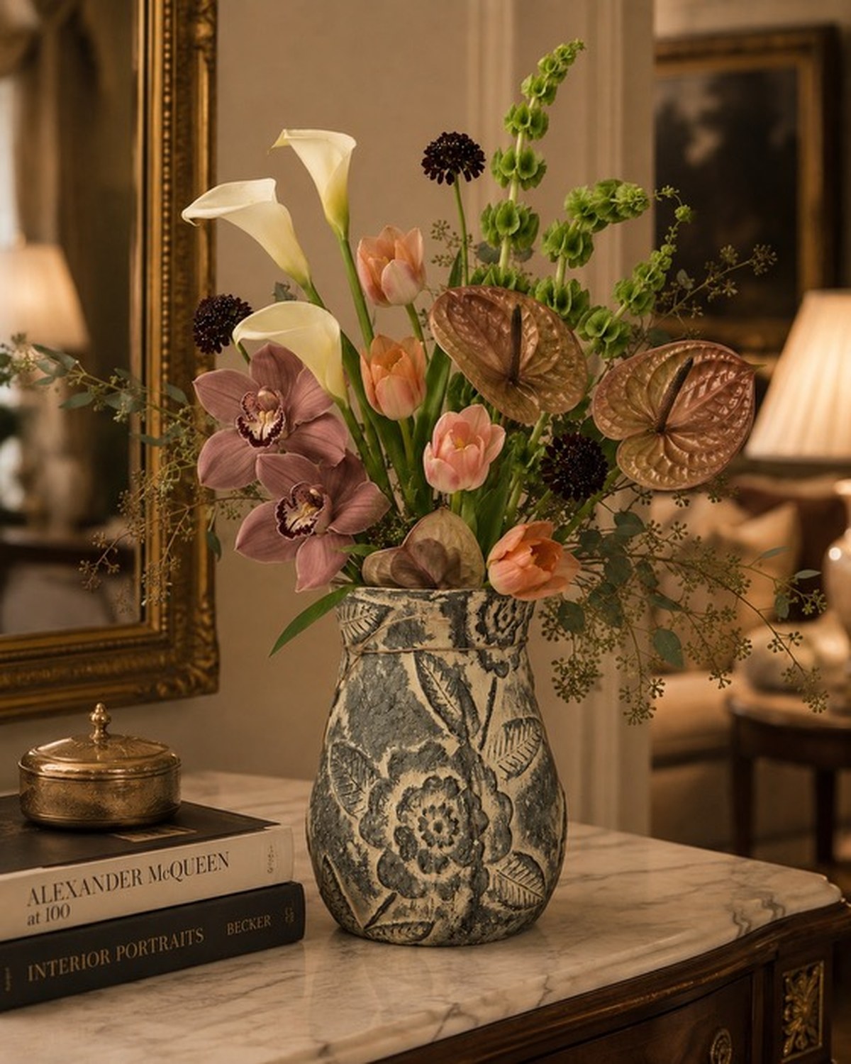 A carved floral stone vase holding mauve orchids, peach tulips, white calla lilies and bells of Ireland beneath a gilded arched mirror.