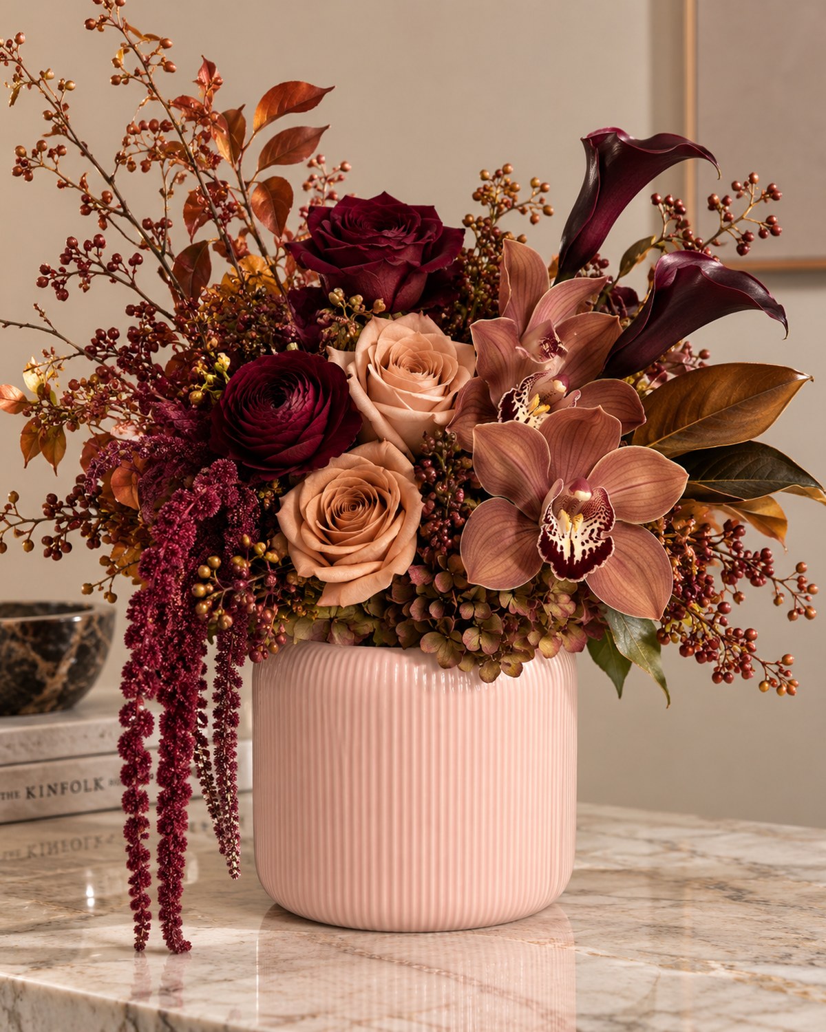 A ribbed blush vase of deep burgundy roses, magnolia leaves and trailing amaranthus in a rich autumn palette.