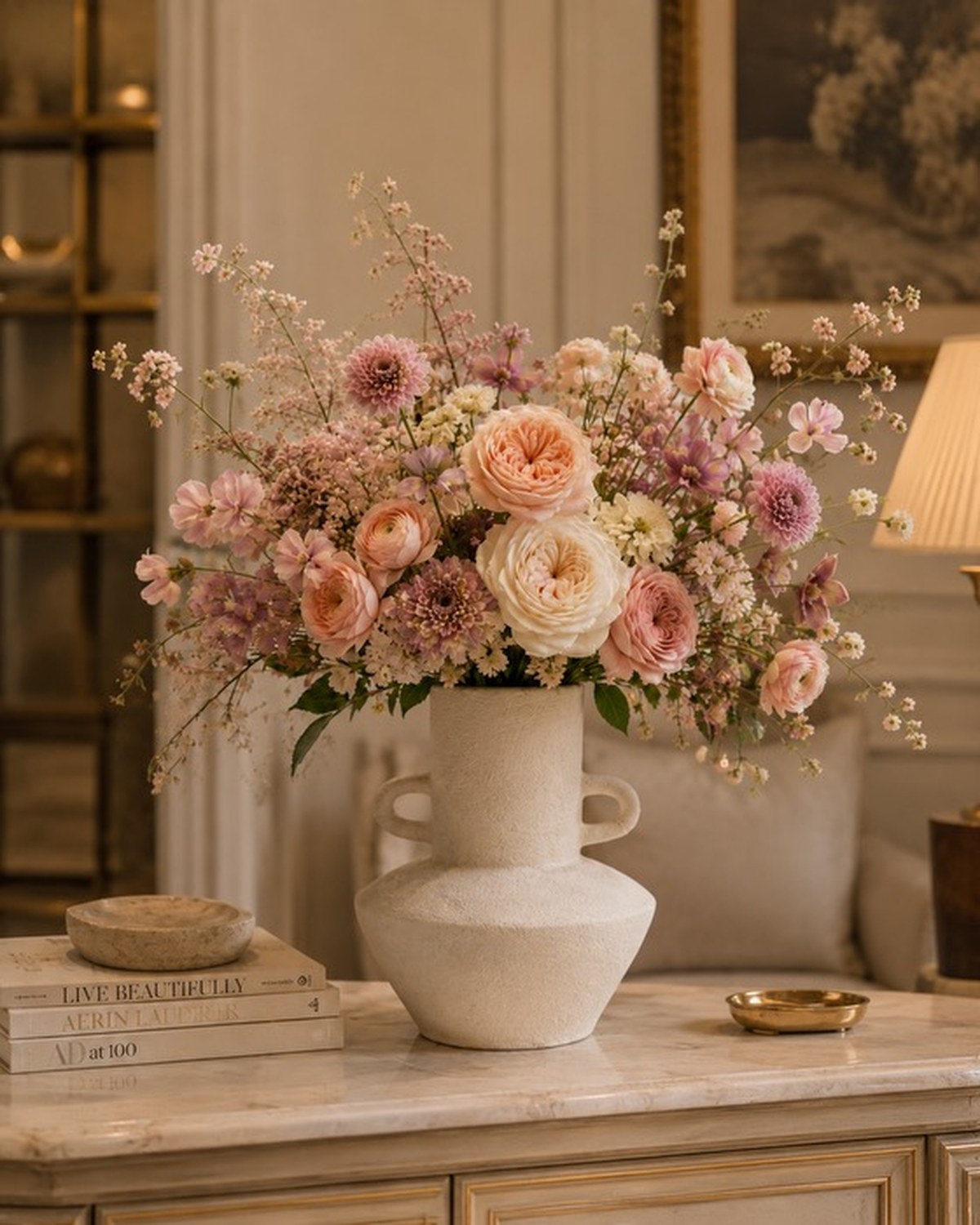 A textured white vase on a marble pedestal with white peonies, snapdragons, mauve roses and Queen Anne&rsquo;s lace.