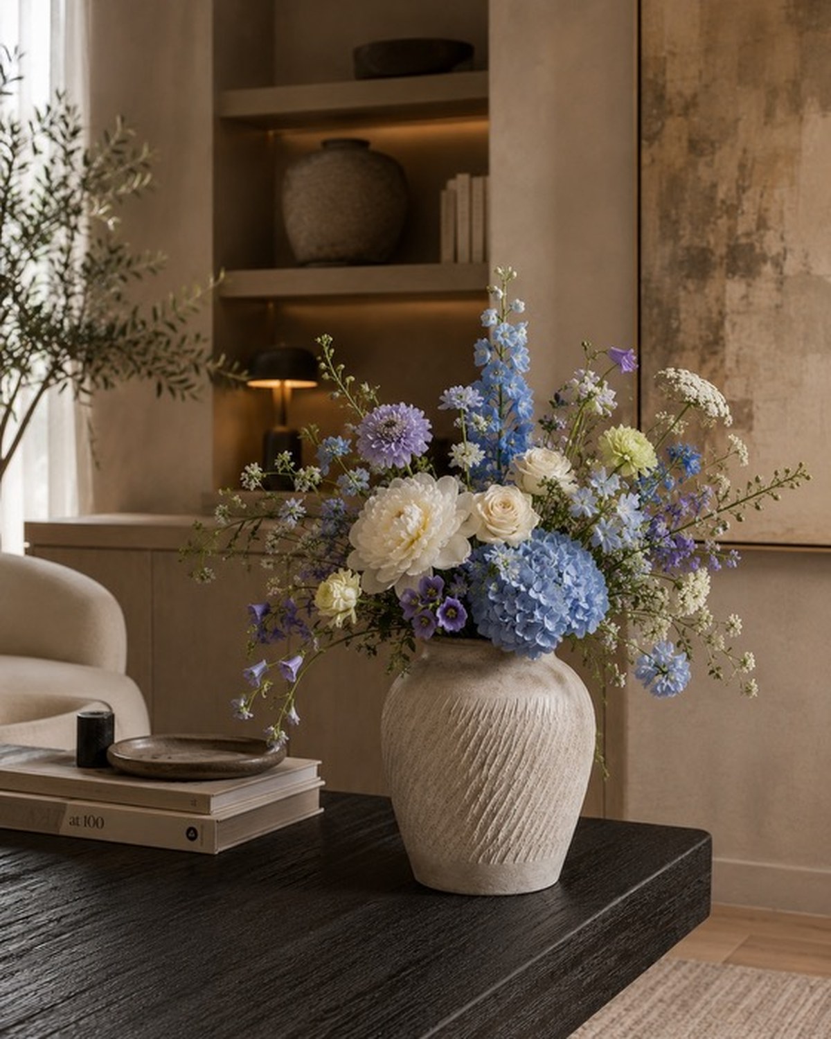A textured stone urn holding blue hydrangea, lavender delphinium and ivory ranunculus on a dark wood table.
