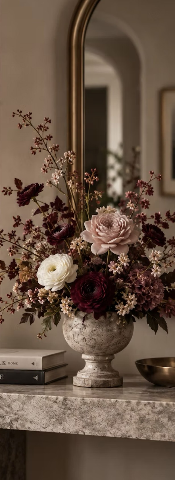 An arched brass mirror above a console holding a stone urn of burgundy, blush and ivory flowers with two stacked books.